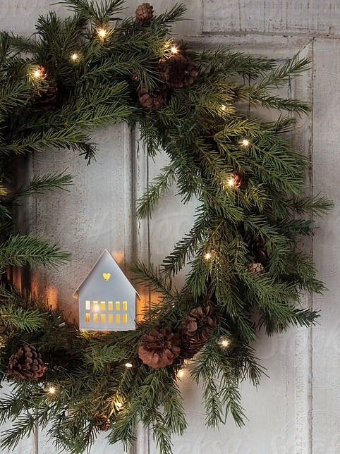 An evergreen holiday wreath with pine cones, lights, and a white house with lights inside is a cozy and cool idea