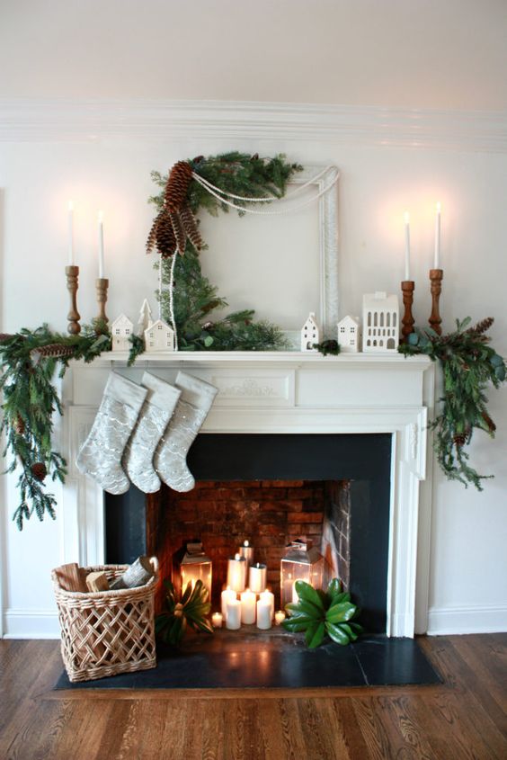 a beautiful Christmas fireplace with pillar candles and candle lanterns, a basket with firewood, evergreen plants and tall, thin candles