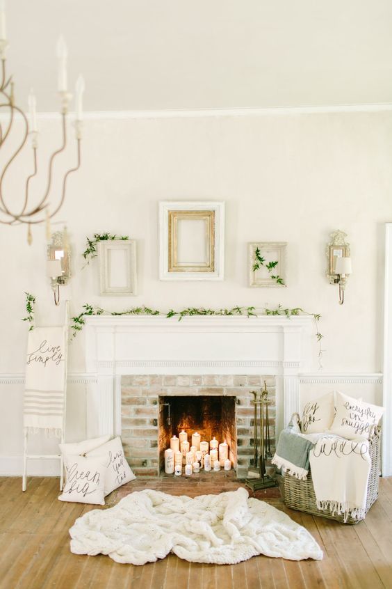 a neutral and cozy room with a fireplace with pillar candles, a rug, a basket with pillows, evergreen plants and empty frames