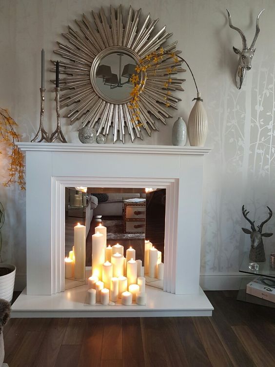 an artificial white fireplace with a mirror shade and pillar candles, a mirror with sunrays and vases on the mantel as decoration
