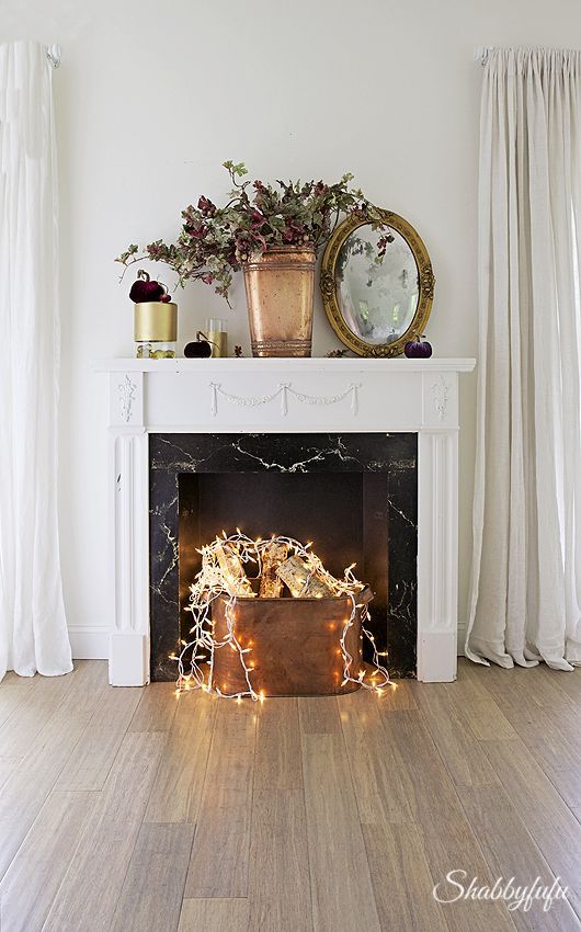a sophisticated non-functioning fireplace in white and with a black marble part, with a copper bucket containing firewood and lights