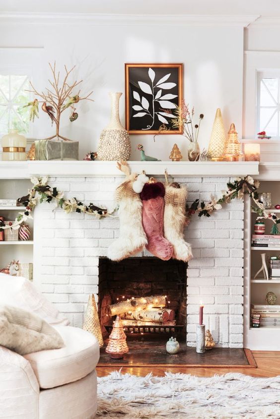 A white brick fireplace with firewood with lights, colorful fur stockings and artificial leaf garlands for Christmas