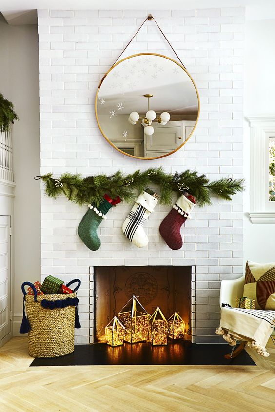 a Christmas fireplace with candle lanterns and lights, evergreens and stockings over the fireplace and a basket of presents