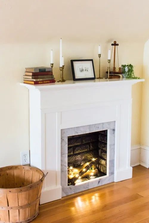 A faux fireplace with logs and lights, as well as a mantel with books, candles, and greenery, is cozy and cool