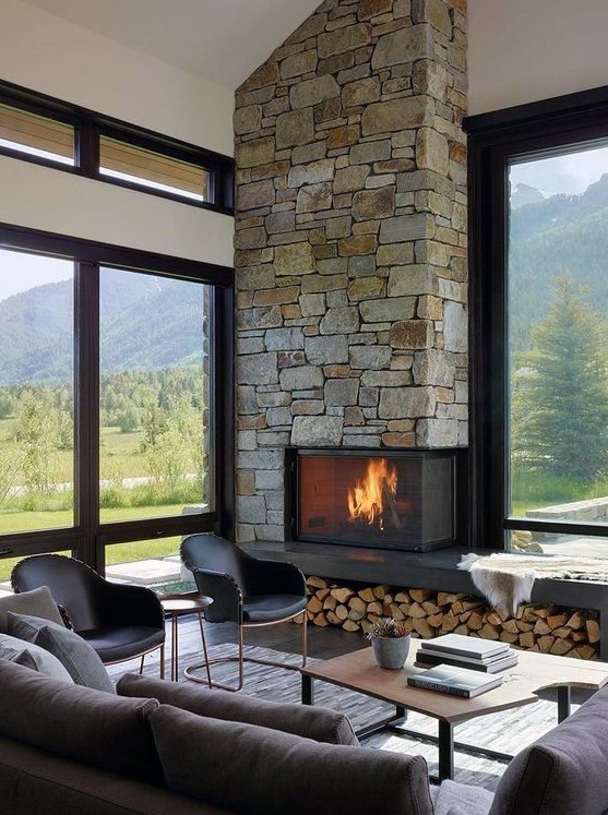 a modern cabin living room with a stone-lined fireplace and firewood storage, chic furniture and lots of windows