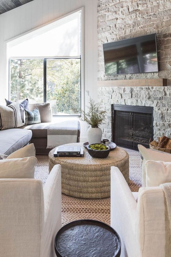 a farmhouse living room with a stone fireplace, a gray sofa, white chairs, a black side table and a jute coffee table