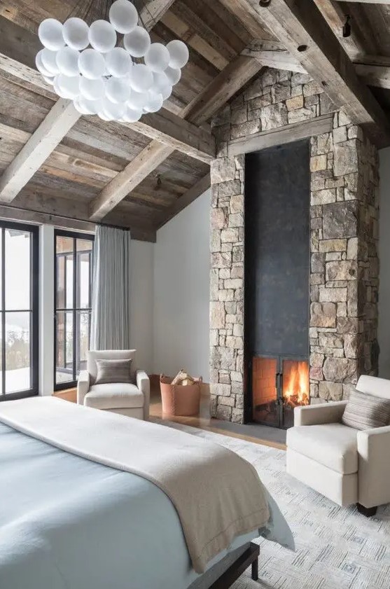 a modern chalet bedroom with a wooden ceiling, a bed with blue linens, a stone-clad fireplace and a bubble chandelier