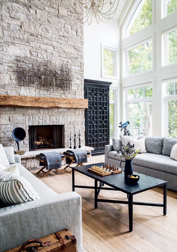 a living room with a stone fireplace, a stained mantel, stools, gray sofas, a black coffee table and unique decor