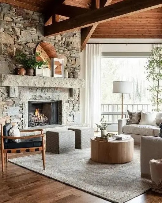 a modern country-style living room with a faux stone-clad fireplace, wooden beams, neutral furniture and a round coffee table