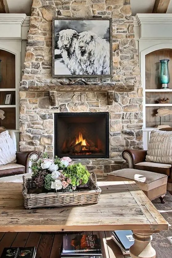 A modern farmhouse living room with a stone-clad fireplace, vintage furniture, a stained coffee table, artwork and flowers