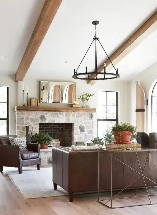 a modern farmhouse living room with a stone-clad fireplace, exposed beams, leather furniture, a metal chandelier and potted plants