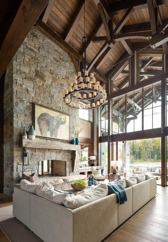 a living room in a mountain cabin with a wooden ceiling, a stone-clad fireplace and stylish, neutral furniture, as well as a statement chandelier