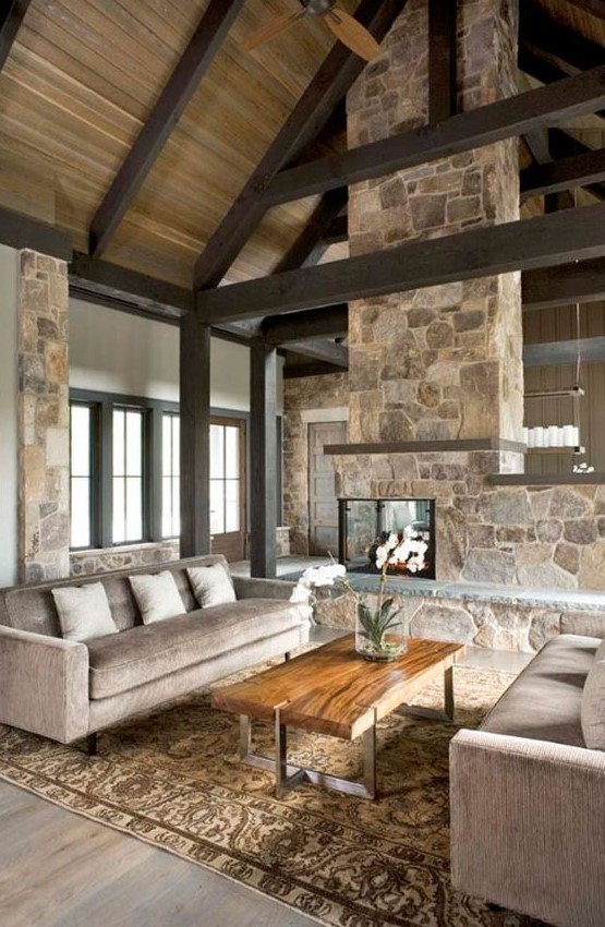 A neutral, modern cabin living room with a double-sided stone fireplace, neutral furniture and exposed beams