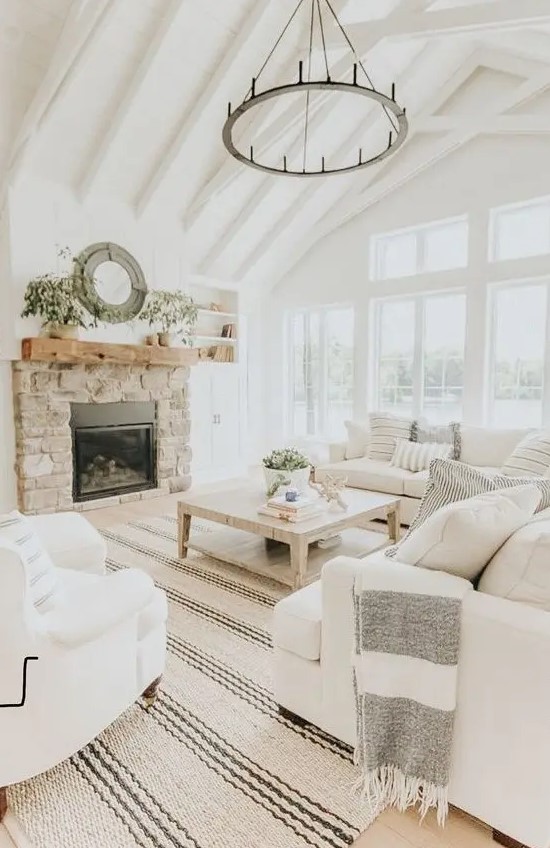 a neutral farmhouse living room with exposed beams, a round chandelier, white furniture and a built-in stone fireplace