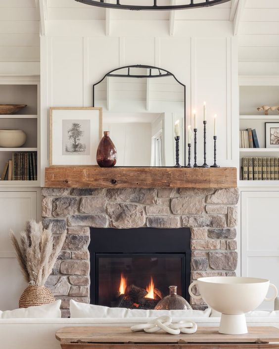 A neutral farmhouse room with a stone-clad fireplace and a wooden mantel, candles, a mirror and some decorations