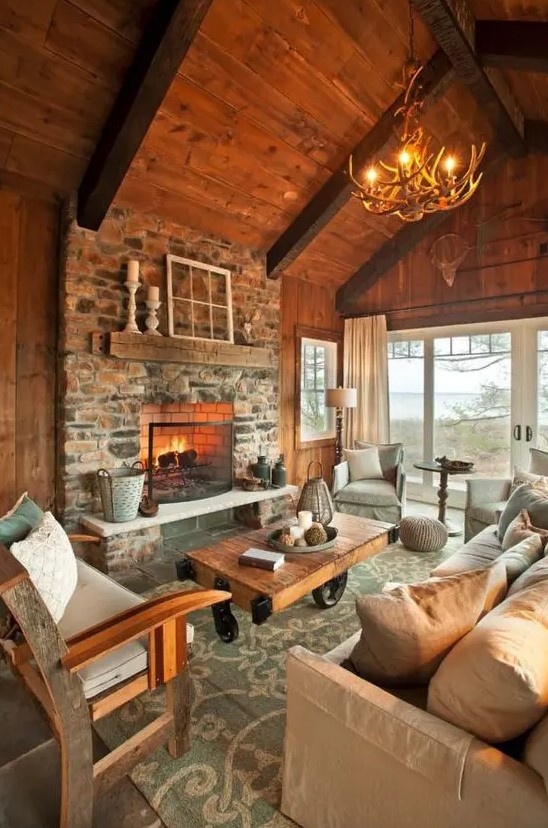 An inviting chalet living room with wooden ceiling and walls, a stone-clad fireplace, modern and rustic furniture and an antler chandelier