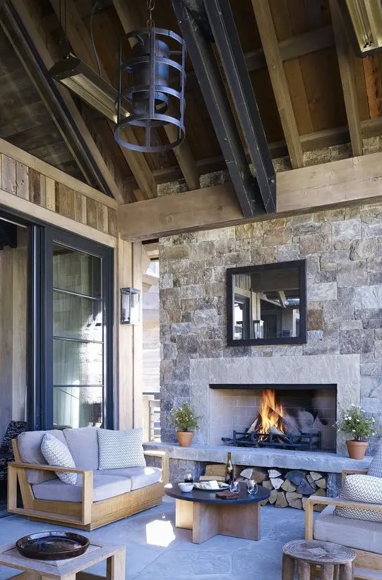 An outdoor space under a roof with a stone fireplace, neutral modern furniture, metal pendant lamps and a low coffee table is chic