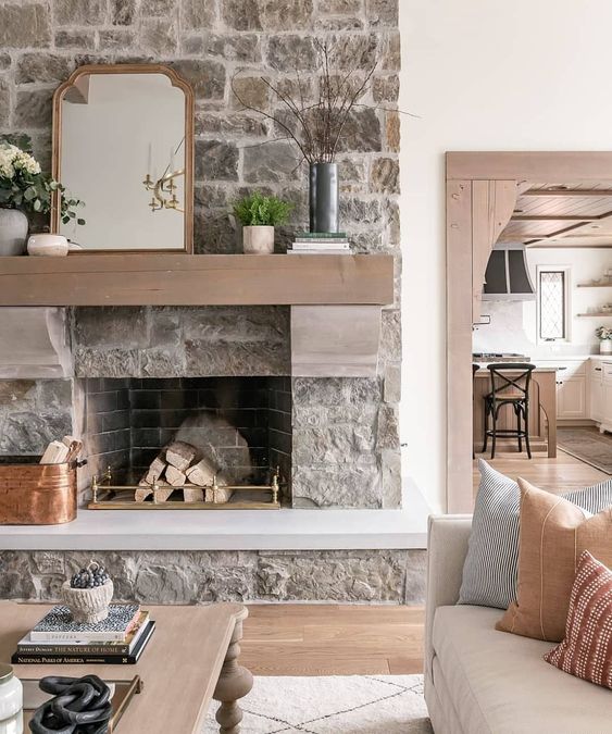 a neutral rough stone fireplace with firewood on a gold stand, a light brown mantel with potted plants and a mirror on top