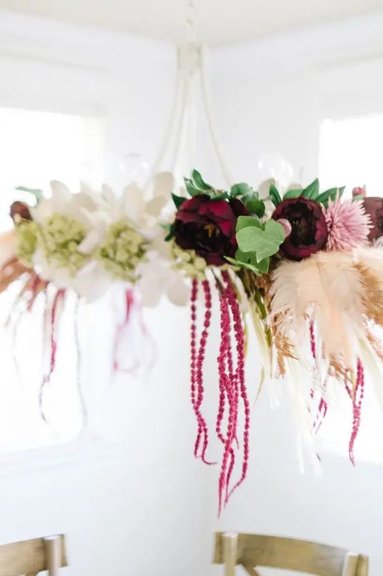 A boho fall chandelier made with burgundy, white and pink flowers and spray painted feathers as decoration