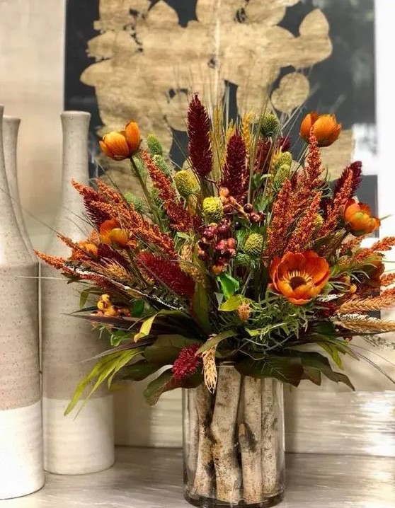 A bright, rustic arrangement of rust, burgundy artificial flowers, dried herbs and green plants and wooden sticks in the vase