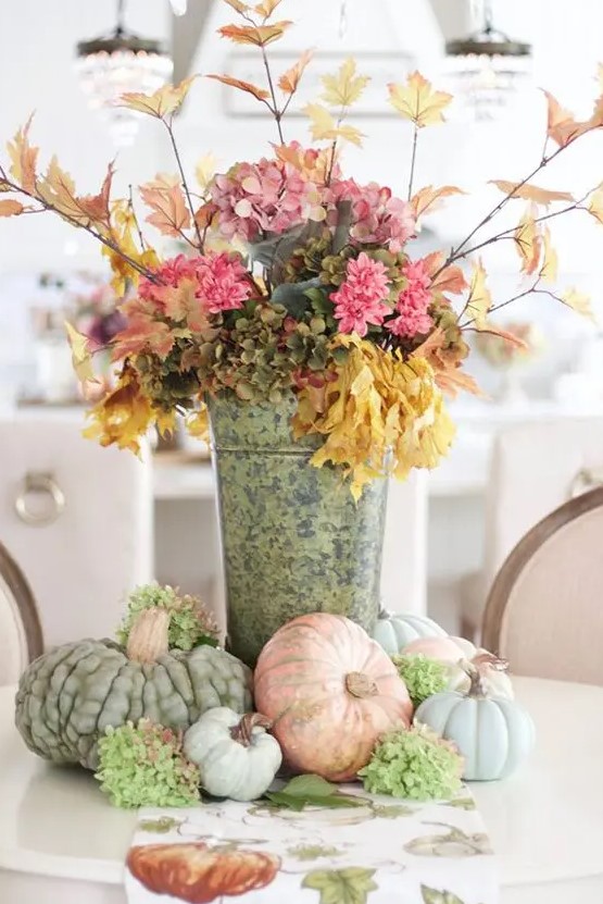A stylish, rustic fall decoration made from a bucket of pink and green faux flowers, branches with leaves and heirloom pumpkins