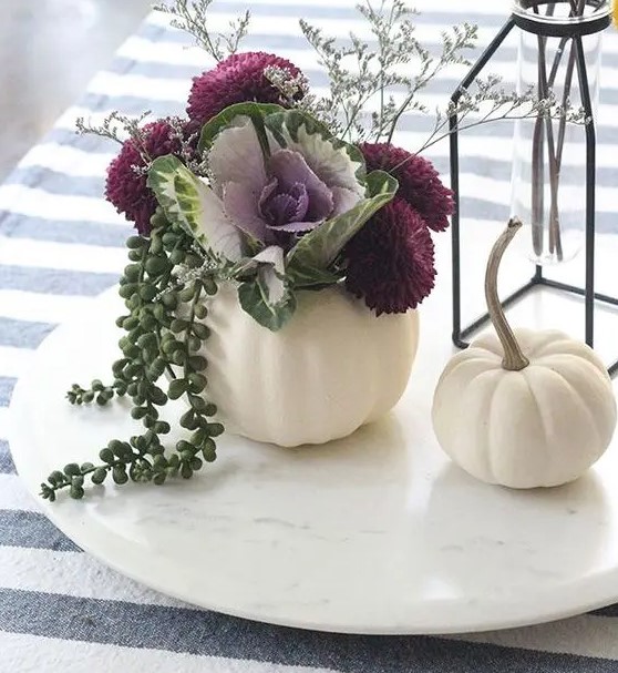 a white pumpkin with deep purple flowers, lots of greenery, a cabbage, grasses and balls in a vase