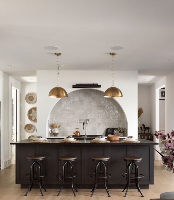 a modern kitchen with an arched niche clad with Zellige tiles and built-in cabinets, a black kitchen island, brass pendant lamps and industrial stools