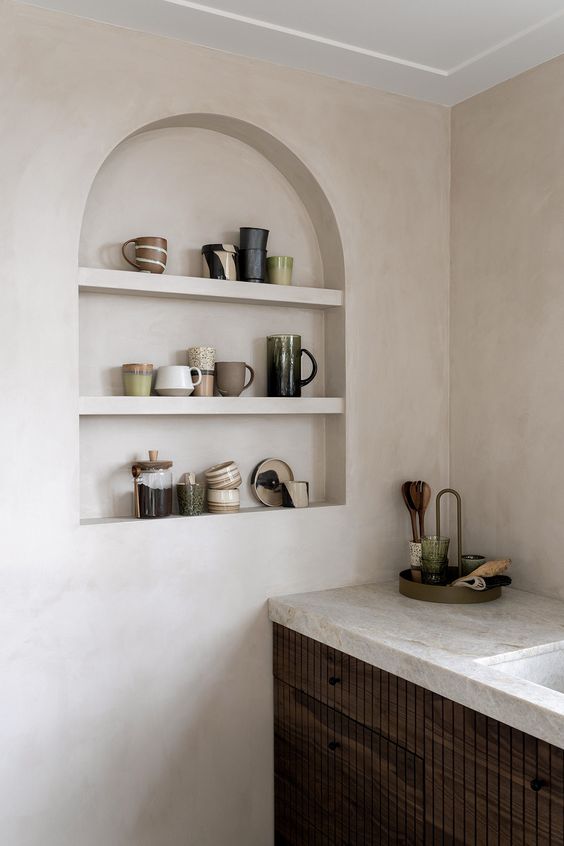 a contemporary kitchen with plaster walls, dark-stained cabines and neutral stone countertops, an arched niche for displaying mugs
