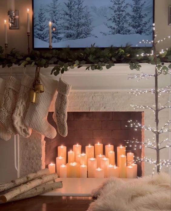 a fireplace with pillar candles, branches, a lighted tree, white stockings and greenery on the mantel