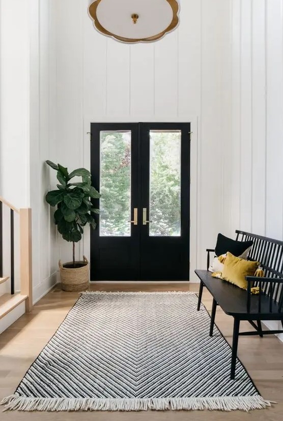 a black and white farmhouse entryway with black doors, a black bench, colorful pillows, a printed rug and a potted plant