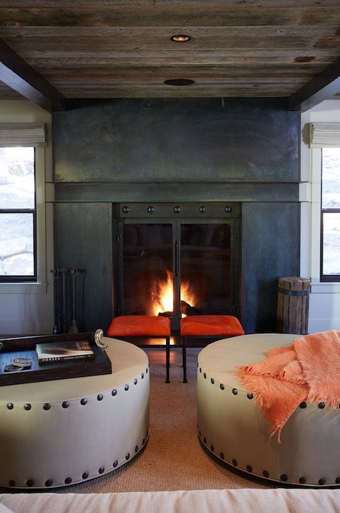 a cozy fireplace nook with a fireplace clad with metal, a duo of orange stools, round ottomans and a reclaimed wood ceiling