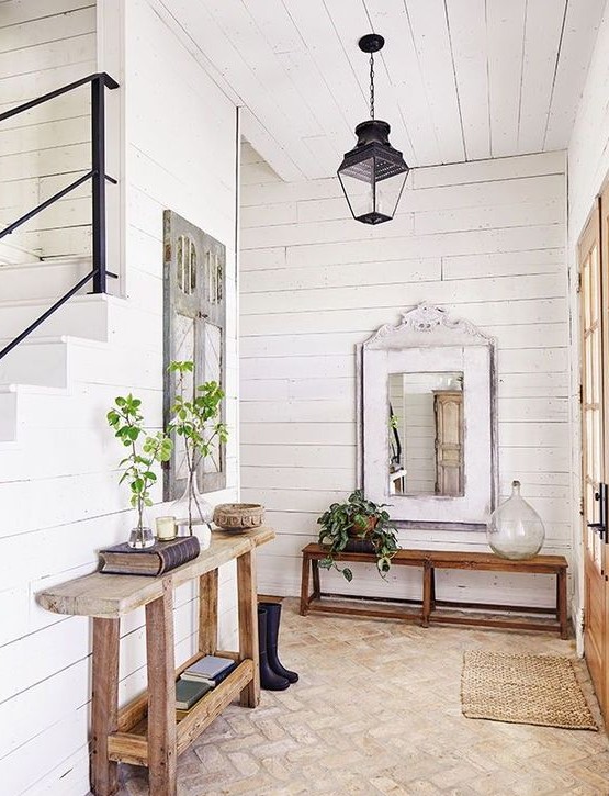 a farmhouse entryway with a console and a bench, a lamp, a jute rug, some greenery in vases and mirrors