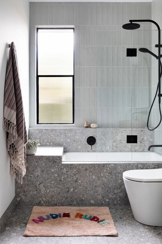 a modern bathroom done with grey terrazzo and grey skinny tile, black fixtures, a colorful rug and towel