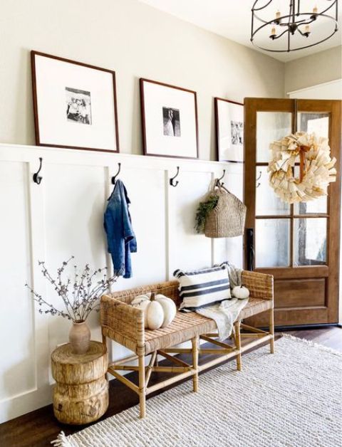 a modern farmhouse entryway with a woven bench, a paneled wall, a side table of wood, a gallery wall and some pumpkins