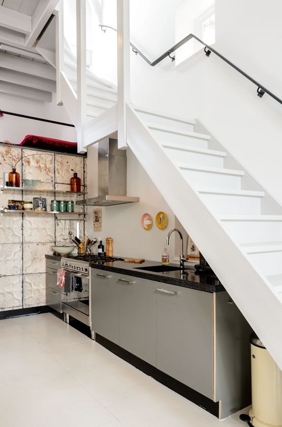 a modern kitchen placed under the stairs, with grey lower row of cabinets, open shelves on the wall and some bright decor