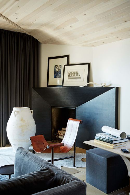 a modern living room with a large metal clad fireplace, a unique chair, a pouf, a coffee table, artwork