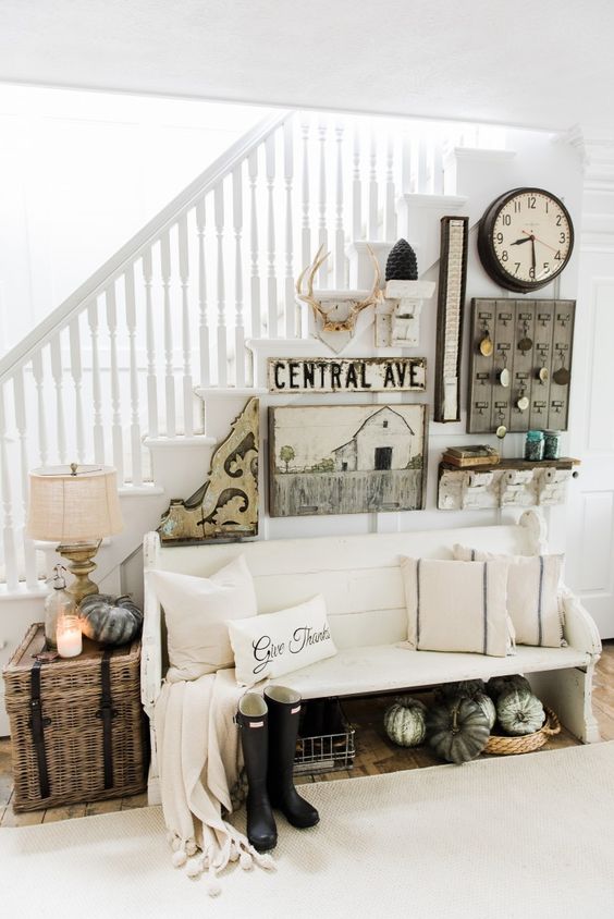 a modern neutral farmhouse entryway with a white bench, a basket chest with a lamp, a gallery wall in rustic style and pumpkins