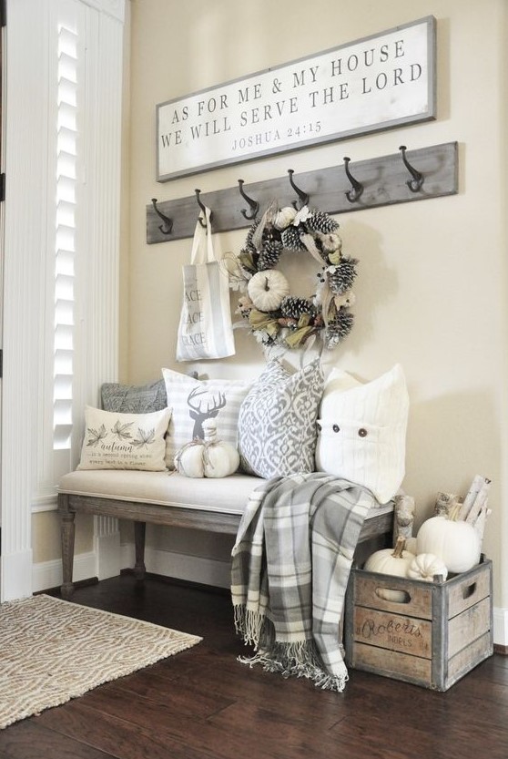 a neutral farmhouse entryway with an upholstered bench, printed pillows, a crate with branches and pumpkins, a rack with hooks and a sign