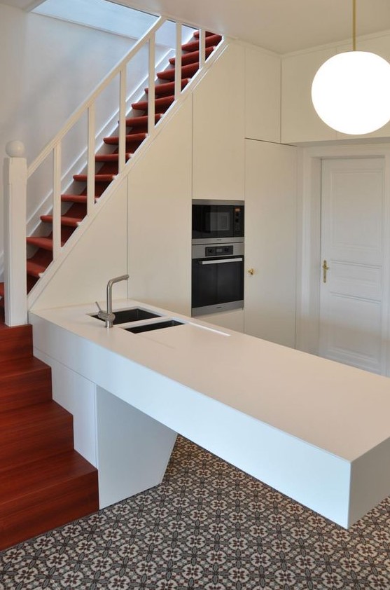 a sleek white kitchen partly built into the staircase, with an architectural kitchen island and a pendant lamp