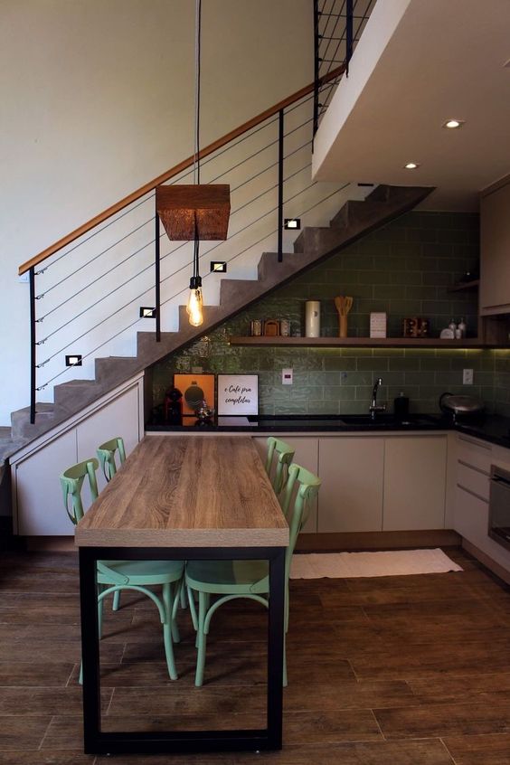a small industrial kitchen with grey cabinets, black countertops, a green subway tile backsplash, a dining zone and pendant lamps