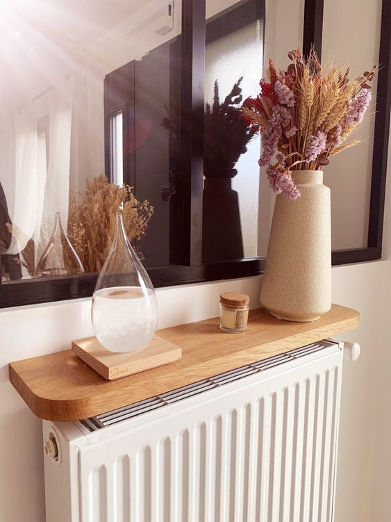a white radiator with a light stained shelf above it, some dried flowers and candles to give the radiator a unified look