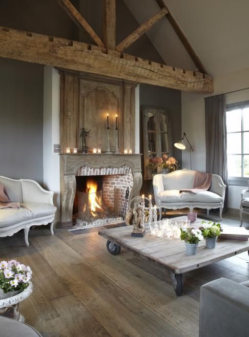 a vintage living room with a brick fireplace, a large wooden beam, gray and cream vintage furniture, light stained floors and a whitewashed coffee table