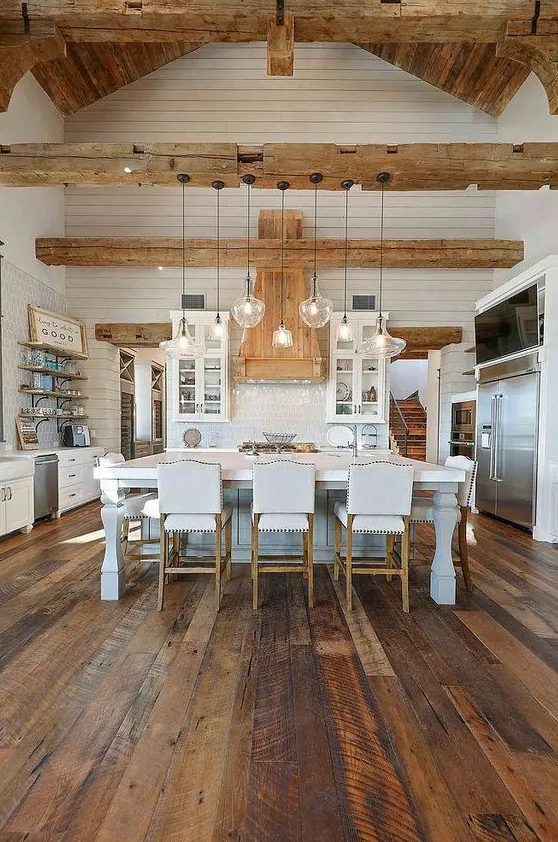 a white farmhouse kitchen with white cabinets, a sophisticated kitchen island, reclaimed hardwood floors, light stained wood beams and a ceiling