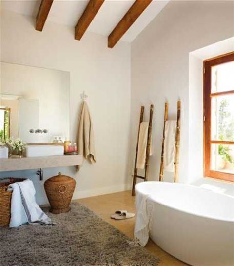 a beautiful coastal bathroom with richly stained wood beams and a window frame, an oval bathtub, a floating vanity, baskets for storage and ladders