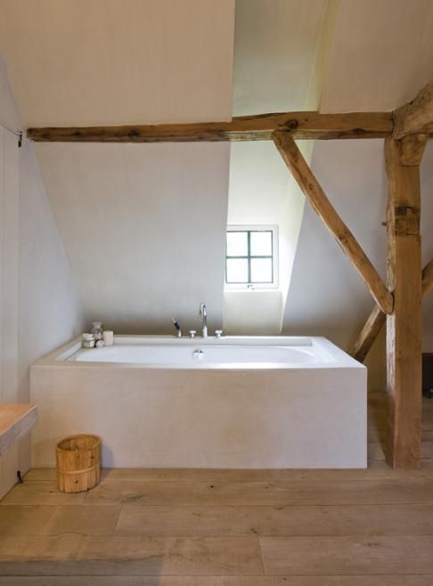 a minimalist, neutral bathroom with a wooden column and beam, a bathtub clad in white stone, a wooden floor and a small window