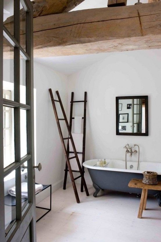 a neutral bathroom with wooden beams, a blue clawfoot bathtub, a bench, dark stained ladders and a black chair and a mirror