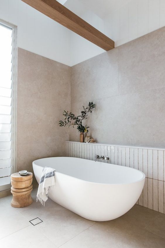 a neutral coastal bathroom with neutral large format and white thin tiles, an oval bathtub, a wooden beam and a wooden stool