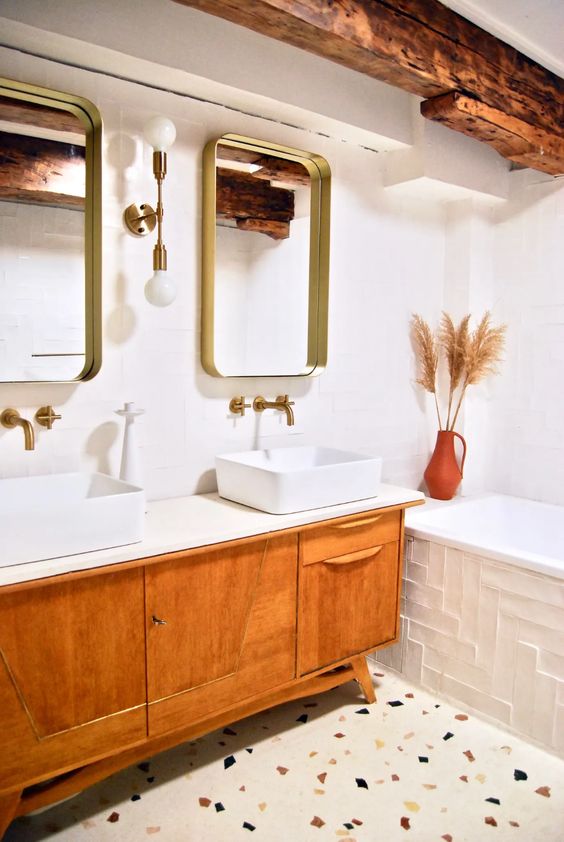 a fairly bright bathroom with terrazzo flooring, a light stained vanity with two sinks, mirrors in gilded frames and a richly stained wooden beam