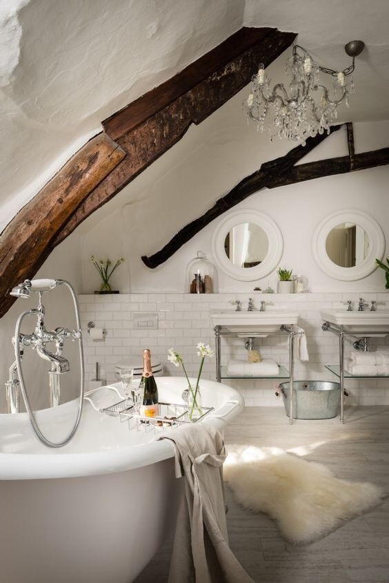 a sophisticated white bathroom with white subway tiles, whitewashed floors, two freestanding sinks, round mirrors and dark stained beams