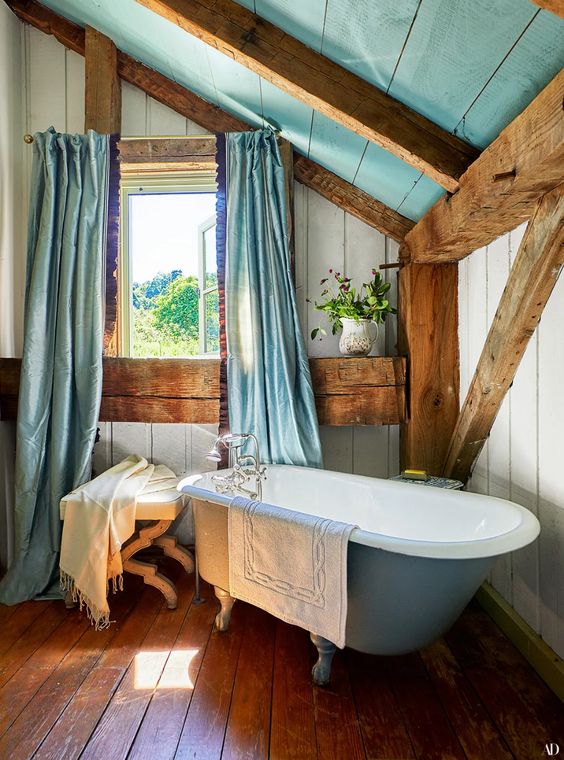 a rustic blue and white bathroom with wooden boards, a blue clawfoot bathtub, lots of wooden beams and columns and blue textiles